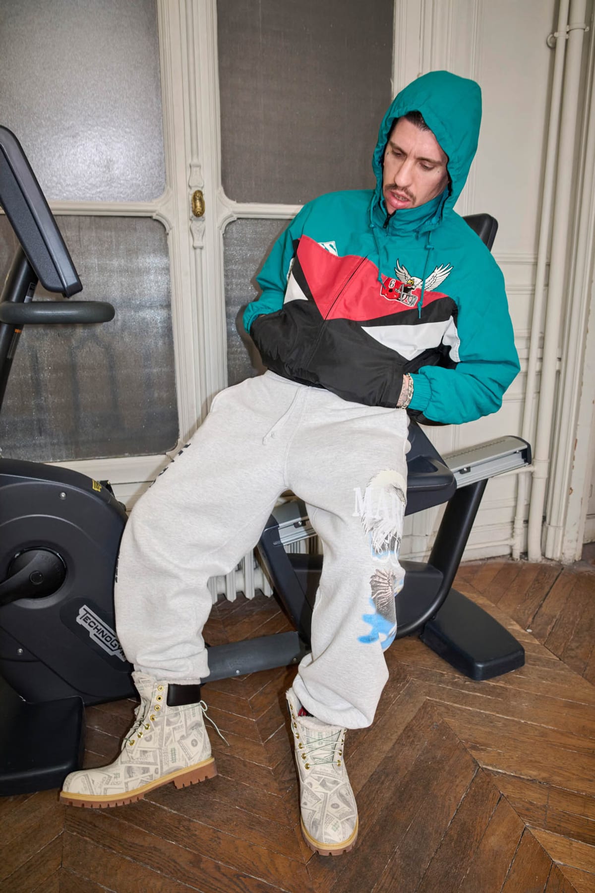 A model on an exercise bike in a hooded teal, red and black anorak with an eagle patch, grey sweatpants and dollar-print Timberland boots, from the Supreme x MM6 Maison Margiela Spring 2026 lookbook